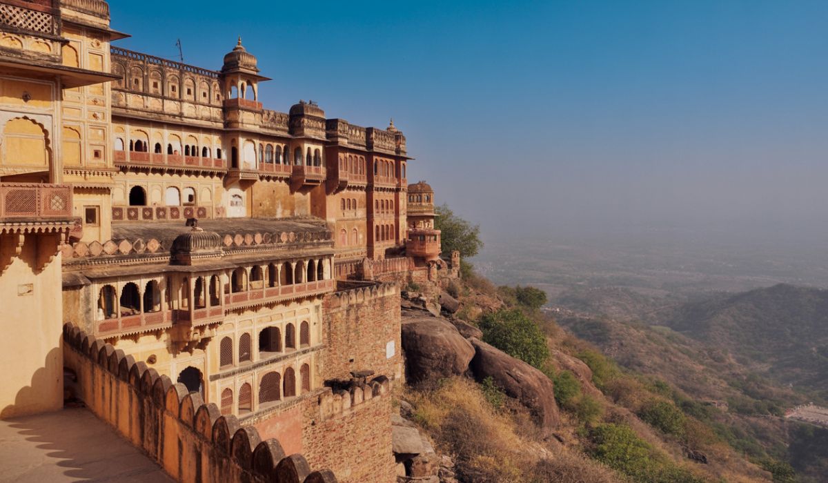 Historic Neemrana Fort-Palace built along a hillside, featuring layered sandstone architecture, arched windows, and balconies overlooking a vast valley and distant landscape under a clear sky.